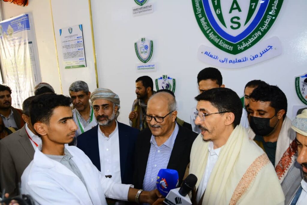 Group of men gathered for a press interview at a science/education event, with microphones held toward a young man in a white coat.