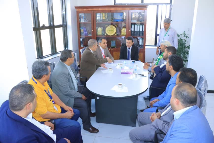 Eleven professionals in suits seated around a conference table, engaged in a meeting, with a trophy cabinet in the background.