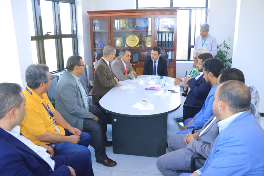 Group of professionals in business attire seated around a round conference table, engaged in a meeting in a bright office.