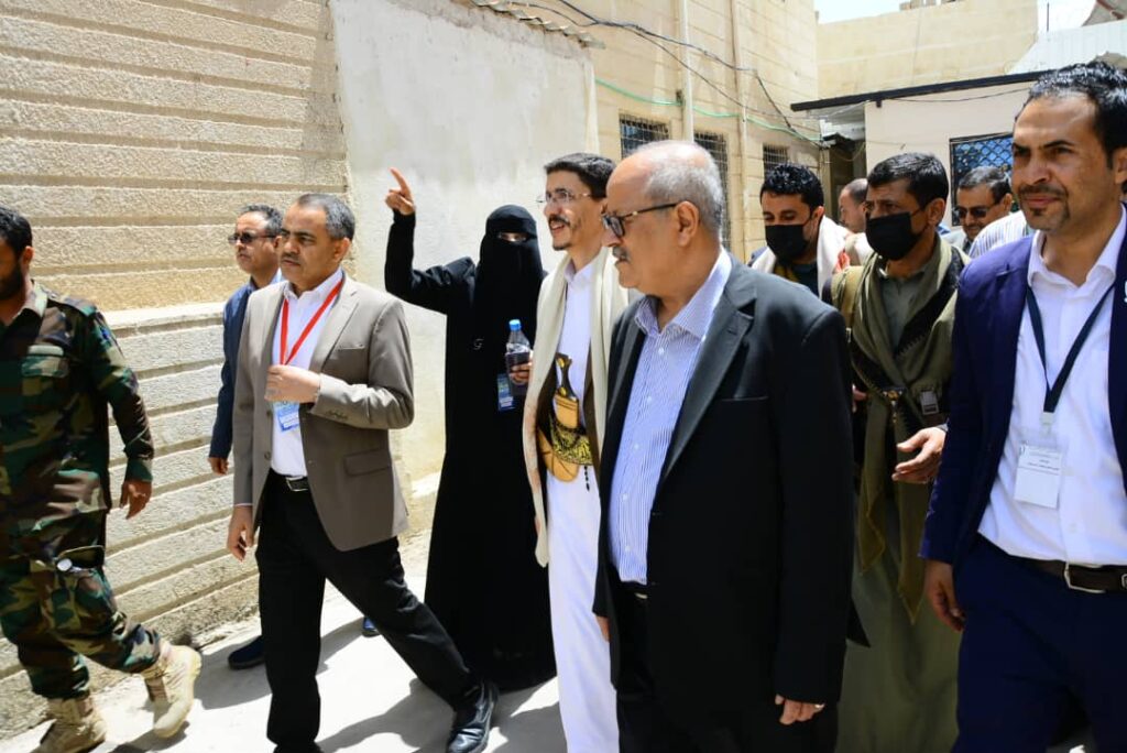 Group of formally dressed officials walking through a sunlit alley, some wearing lanyards and badges, with a woman in a niqab among them.