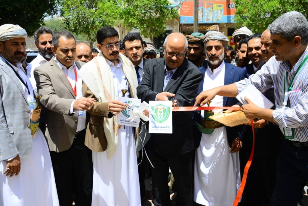 Group of men in traditional clothing and suits cut a red ribbon at an outdoor ceremony.