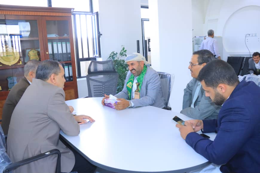 Five men sit around a white oval conference table in a bright office; one man with a green scarf and head covering holds a tissue box while others listen.