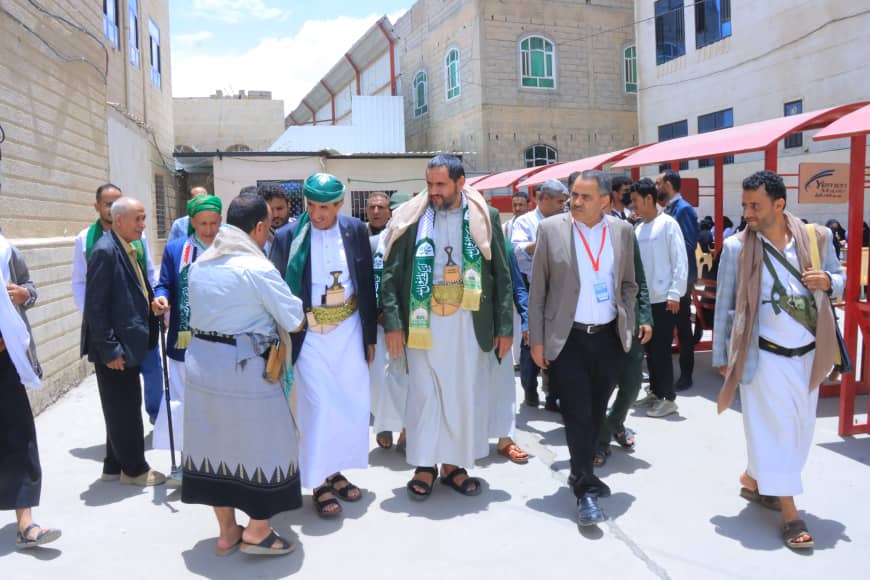 Group of men in traditional white robes with green sashes gather in a sunlit courtyard with red umbrellas and beige buildings in the background.