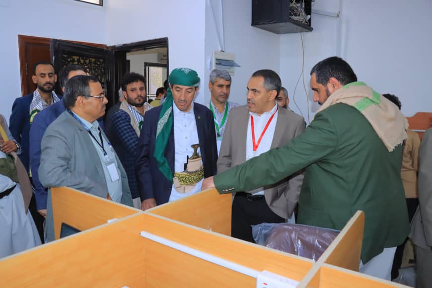 Group of men in a workshop examining wooden partitions or desks during a tour; one man wears a green cap and traditional belt, others in suits observe.