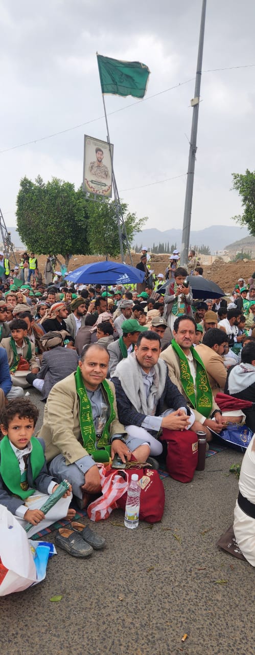 Crowd of men and children sitting on a road at an outdoor gathering, many wearing green scarves; a green flag and a poster pole loom overhead.
