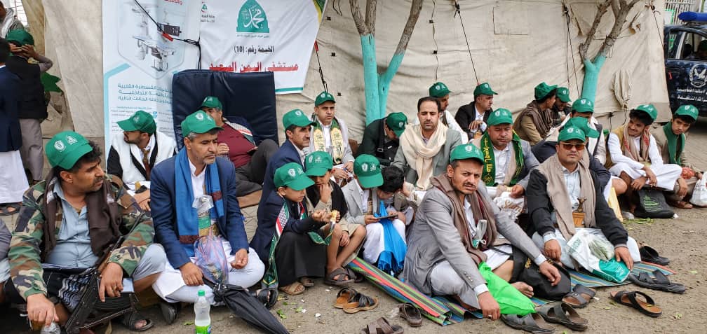 Group of men sitting on the ground in a tented area, many wearing green caps and traditional dress, listening or waiting for an event.