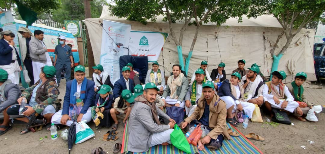 Group of men in green caps sitting on a striped mat outside a large tent at a relief or charity event, with banners in the background and bags of supplies nearby.