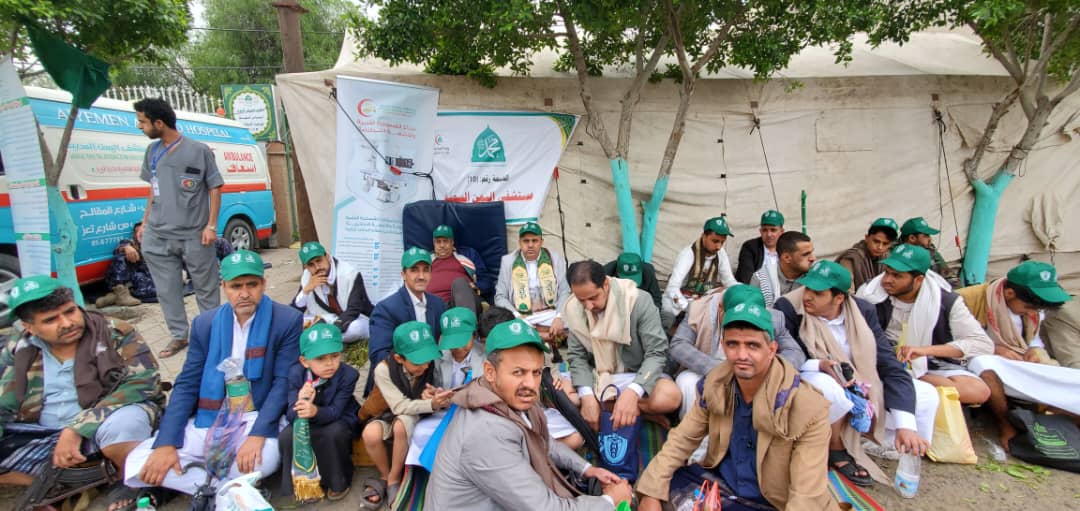 Group of men wearing green caps seated together at an outdoor relief event, with banners and a van in the background.