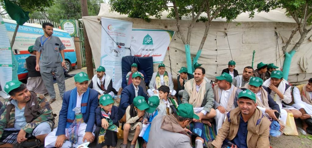 Group of men and boys wearing green caps sit together outdoors at a relief camp with banners behind them.