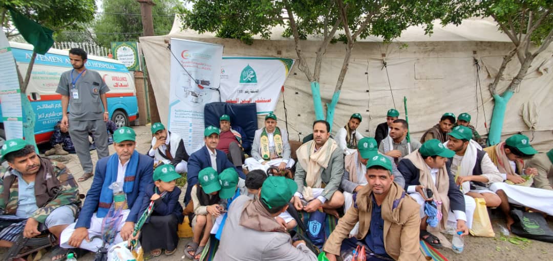 Group of men and boys in green caps sit on the ground at an outdoor relief camp with banners in the background.