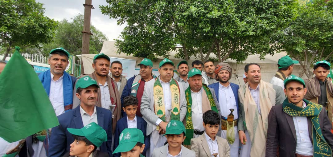 Group of men and children wearing green hats and scarves posing outdoors at a community event in a park-like setting with trees behind them.