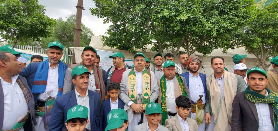 Group of men and boys wearing green caps posing for a photo at an outdoor ceremony with trees in the background.