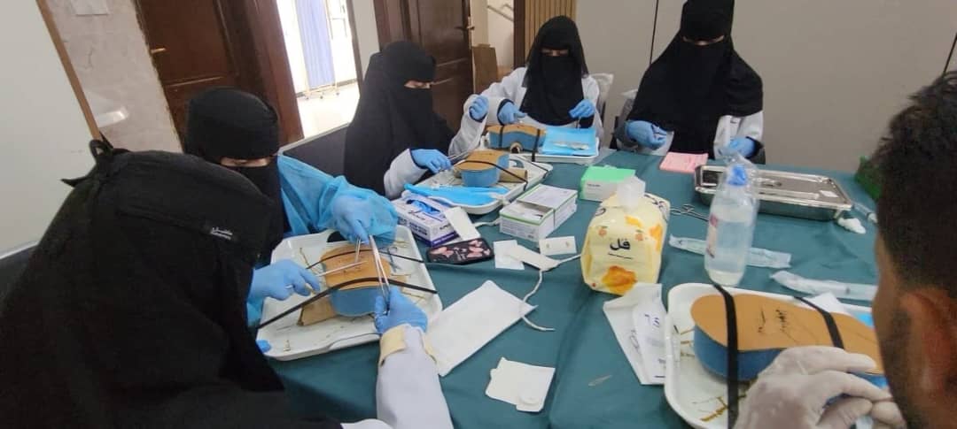 Five women in hijabs wearing gloves and masks practice dental procedures on models around a table in a clinic-like room.