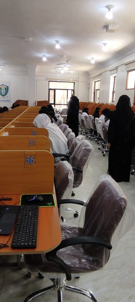Computer lab scene with students in white hijabs seated at numbered desks (36–38); foreground shows a laptop and mouse on the desk in view of the camera.