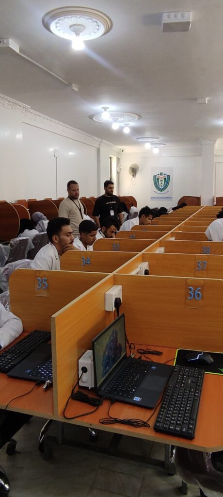 Students in a computer lab seated at numbered cubicles with laptops; two supervisors stand at the back near a banner.