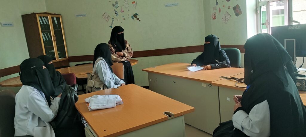 Women wearing niqab and hijab sit around wooden desks in a classroom, a woman at the front holds papers and talks to them, a Dell monitor is visible in the background