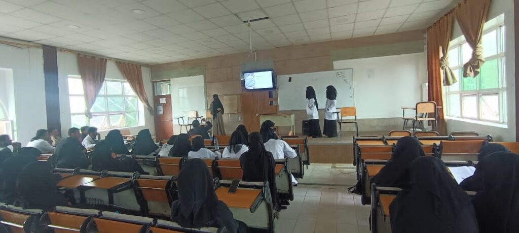 Classroom lecture: two presenters in white coats talk at the front near a whiteboard and a hanging monitor, while students sit at desks watching.