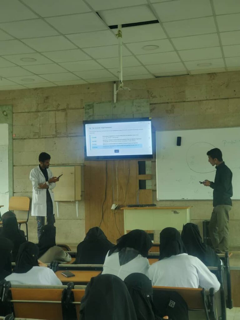 Two presenters at the front of a classroom give a lecture while students in hijabs sit at desks facing them; a monitor displays slides and a whiteboard with a circular diagram is nearby.