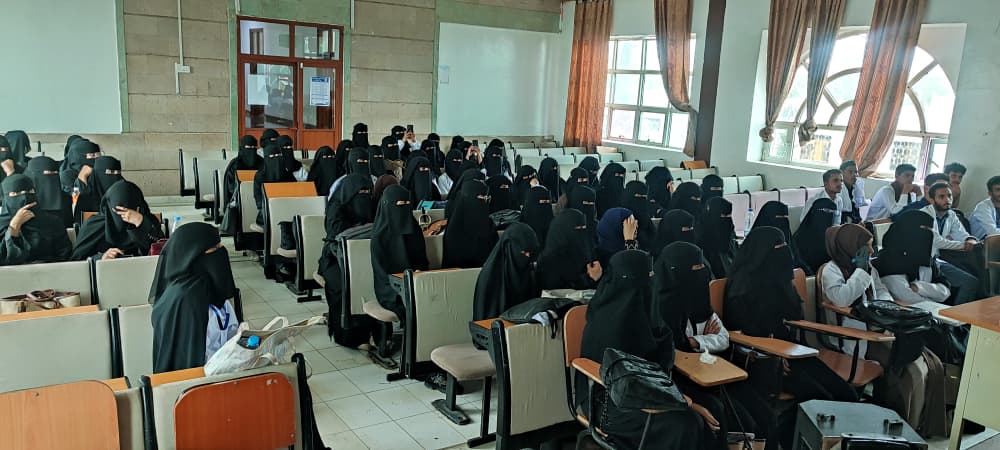 IMG-20260810-WA0004 - جامعة 21 سبتمبر Large classroom filled with students in black hijabs and abayas seated at desks, listening to a lecture; some men sit on the right side.