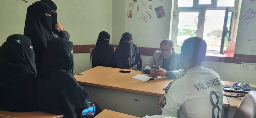 IMG-20260810-WA0006 - جامعة 21 سبتمبر Group of veiled women seated around a table in a classroom as a man shows something on a smartphone.
