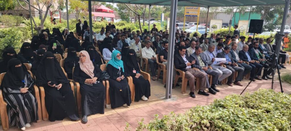 IMG-20260812-WA0000 - جامعة 21 سبتمبر Outdoor event with a large audience seated under a canopy; women in black abayas on the left and men in suits on the right, greenery in the background.