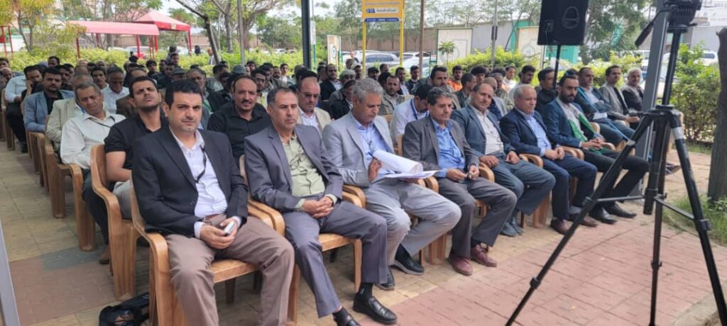 IMG-20260812-WA0001 - جامعة 21 سبتمبر Large group of men in suits seated outdoors at a conference, facing a stage with a tripod camera in front.