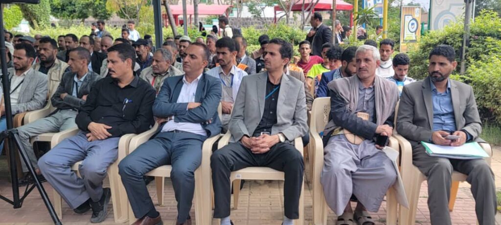 IMG-20260812-WA0003 - جامعة 21 سبتمبر Front row of men seated in plastic chairs at an outdoor event, with a crowd behind them and greenery around.