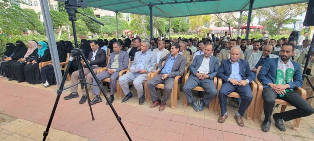 IMG-20260812-WA0004 - جامعة 21 سبتمبر A row of formally dressed men and women seated under a green canopy at an outdoor event, microphone and camera tripod in the foreground.