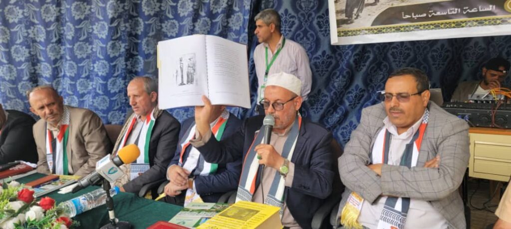 IMG-20260812-WA0014 - جامعة 21 سبتمبر Man in a white cap raises an open book and speaks into a microphone at a seated panel of officials.