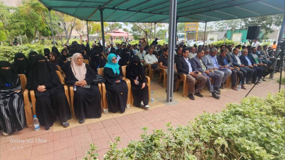 IMG-20260812-WA0015 - جامعة 21 سبتمبر Outdoor ceremony with rows of seated attendees under a green canopy; many women in black abayas and some in colorful scarves, men in suits nearby.