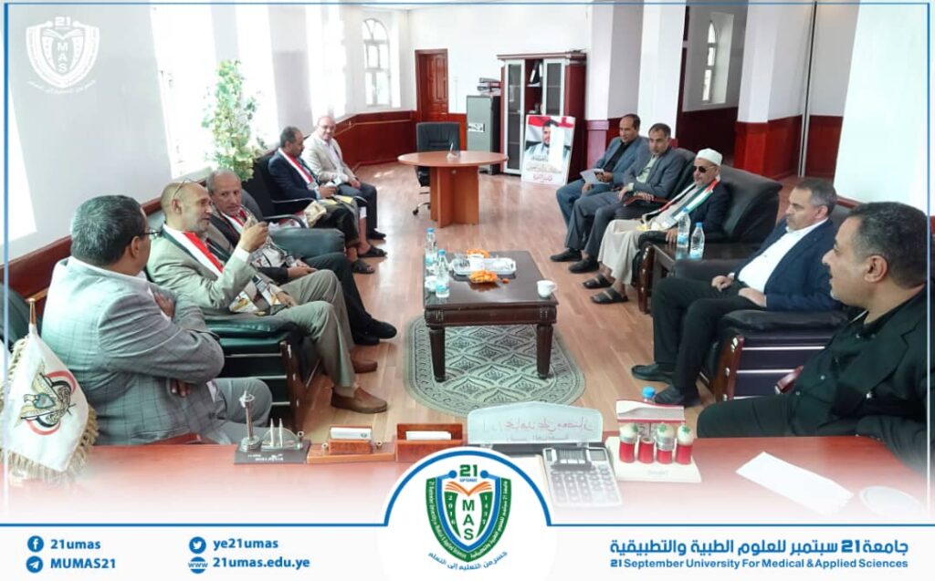 IMG-20260812-WA0016 - جامعة 21 سبتمبر Group of men in suits seated in a semicircle around a coffee table in a university office; banners and the 21 September University logo are visible in the background.