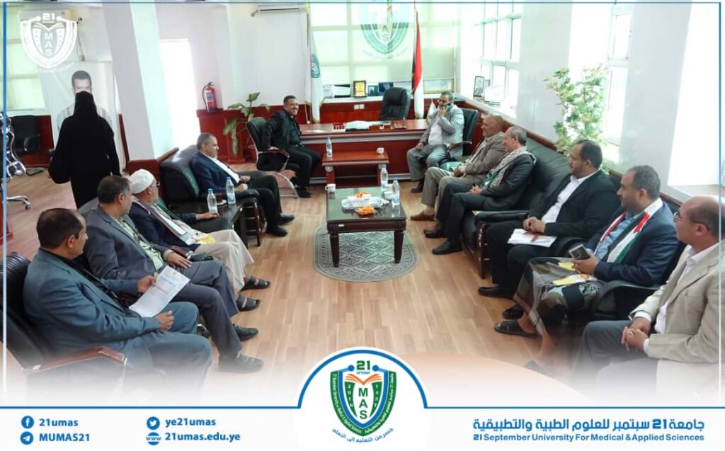 IMG-20260812-WA0018 - جامعة 21 سبتمبر Group of men in suits seated in a U-shaped conference room around a central table, discussing in a formal meeting at the 21 September University of Medical & Applied Sciences.