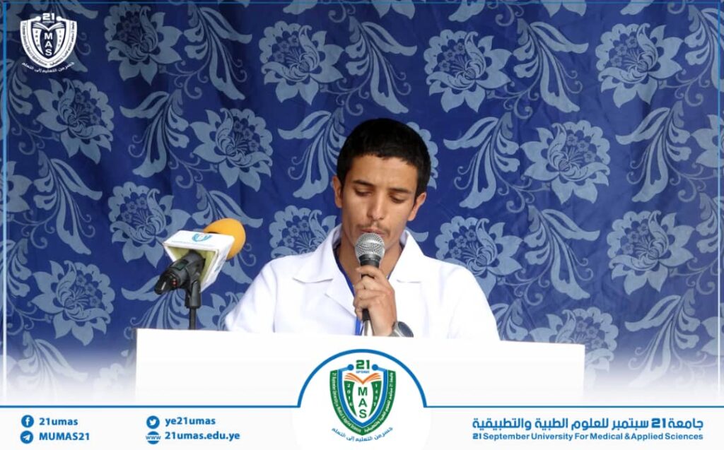 IMG-20260812-WA0020 - جامعة 21 سبتمبر Man in a white lab coat speaks into a microphone at a white podium with university crest, blue floral backdrop behind him.