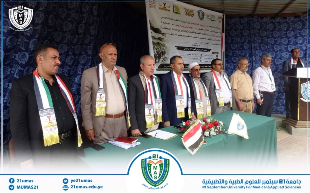 IMG-20260812-WA0021 - جامعة 21 سبتمبر Group of men on a stage at a formal ceremony, wearing sashes and standing behind a green table with flags and flowers.