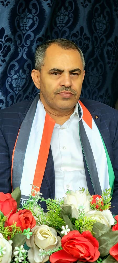 IMG-20260812-WA0023 - جامعة 21 سبتمبر Middle-aged man in a dark pinstripe suit with a tricolor scarf seated at a table with red and white roses against a blue patterned backdrop.