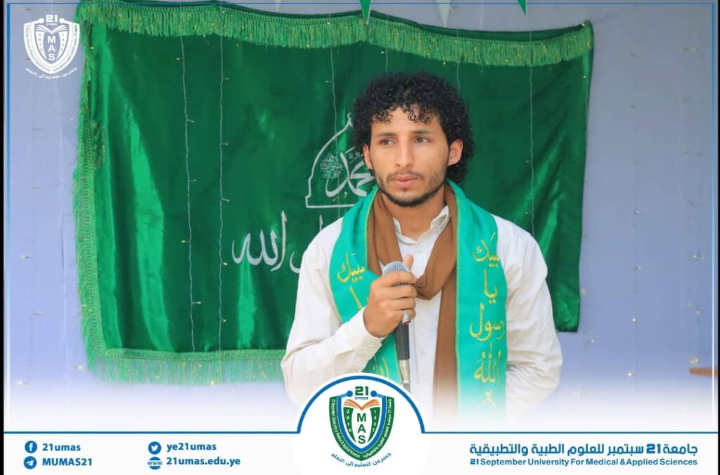 Young man in a white shirt and brown scarf speaks into a microphone in front of a green backdrop with Arabic writing and a university crest visible below.