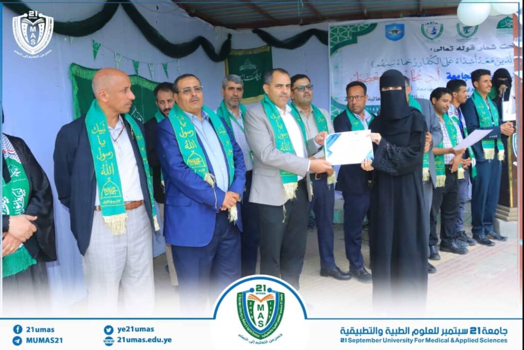 Group of officials on a stage during a certificate award ceremony at 21mas, with men wearing green scarves and a woman in a black abaya receiving a document.