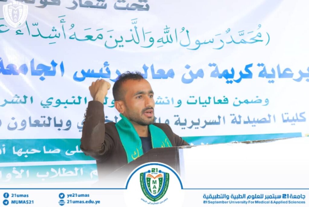 Man delivering a speech at a podium during a university event, with a large Arabic banner behind him.
