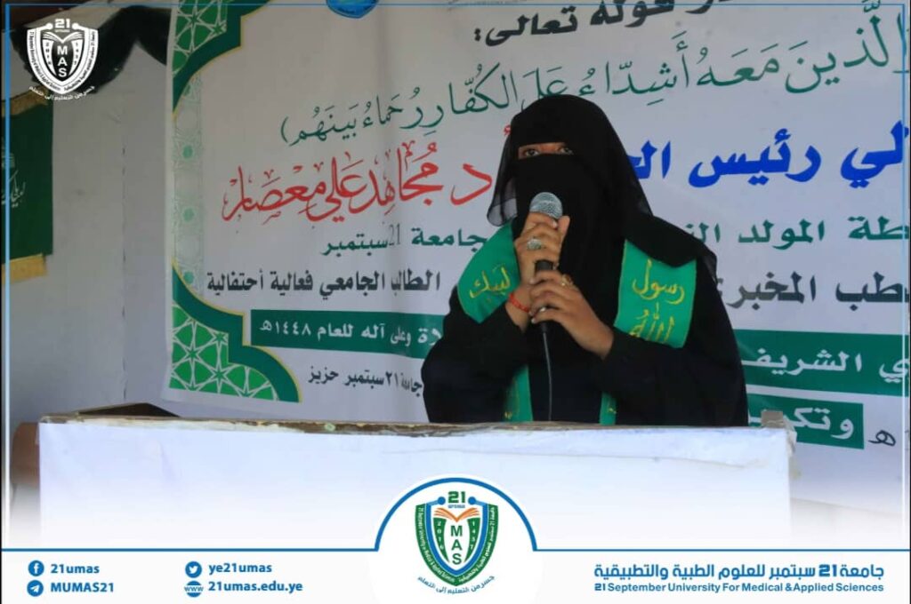 Woman in a black niqab speaks into a microphone at a podium with a large Arabic banner behind her at a university event.