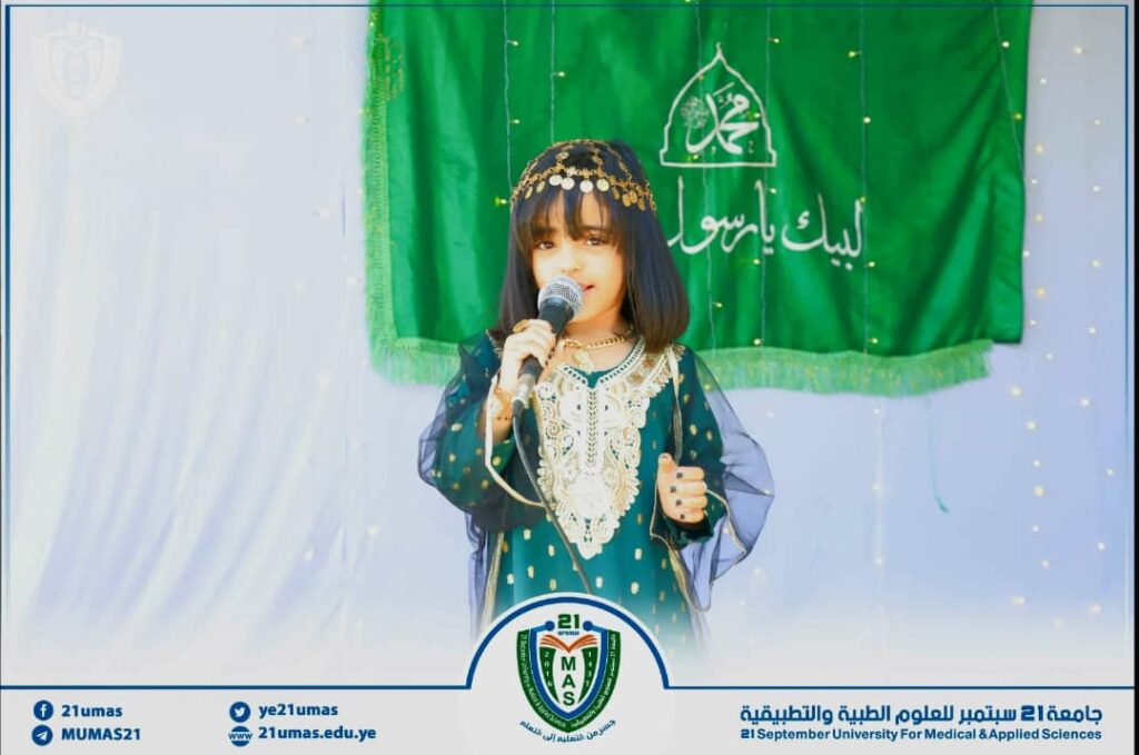 Young girl in a green embroidered dress sings into a microphone on a stage with a green banner behind her.