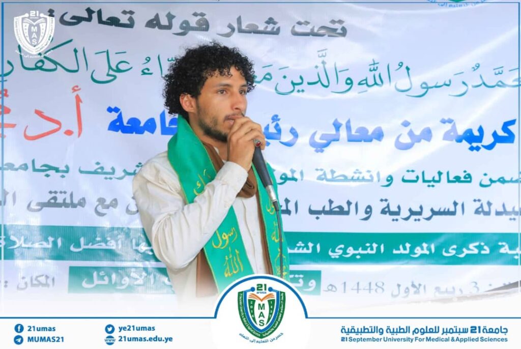 Young man with curly hair speaks into a microphone on a stage, wearing a green scarf; a banner with Arabic text and a university logo is in the background.