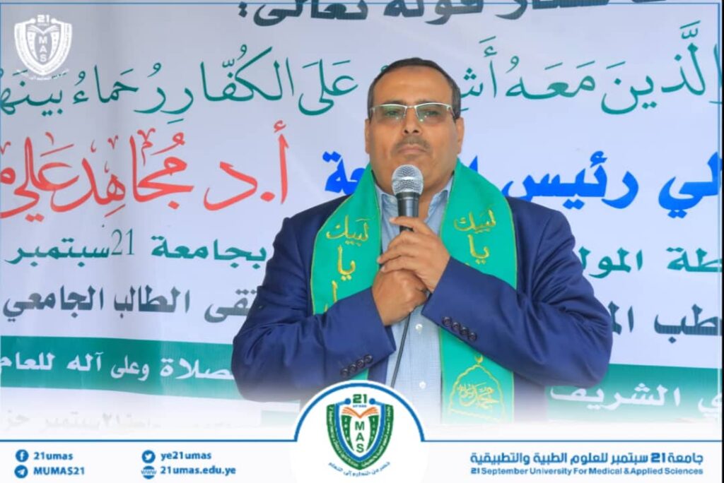 Man in a blue suit with a green scarf speaks into a microphone at a university event banner with Arabic text and a circular logo in the foreground.
