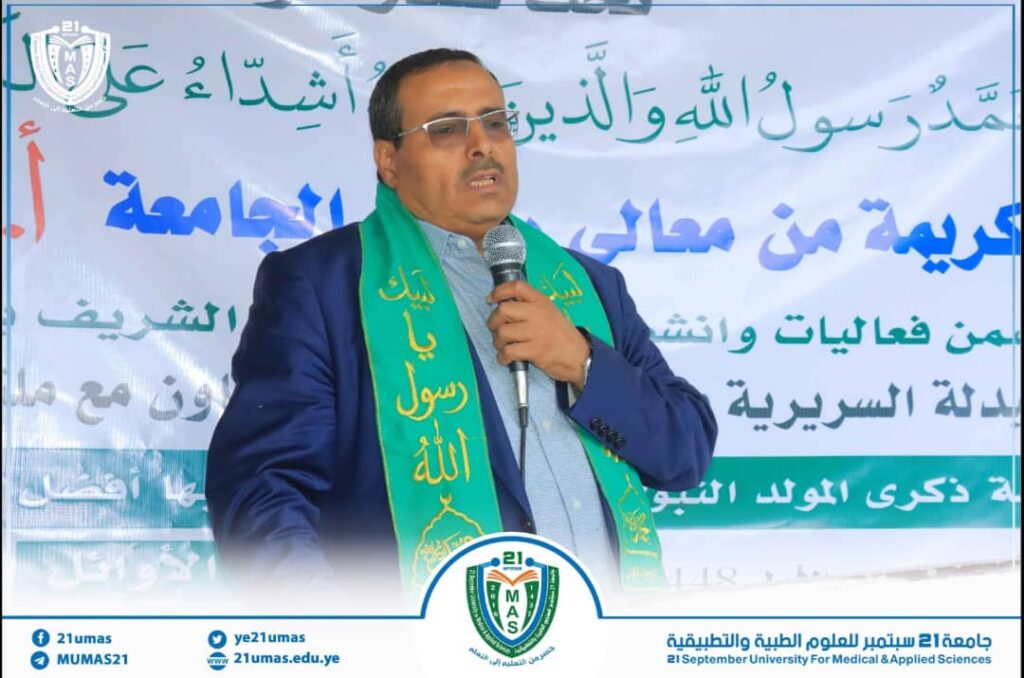 Man in a blue suit with a green scarf speaks into a microphone at a formal event, with a banner and university emblem in the background.