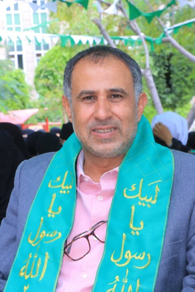 Man outdoors wearing a pink shirt and blue blazer, with a teal scarf that has gold Arabic writing, smiling at the camera.