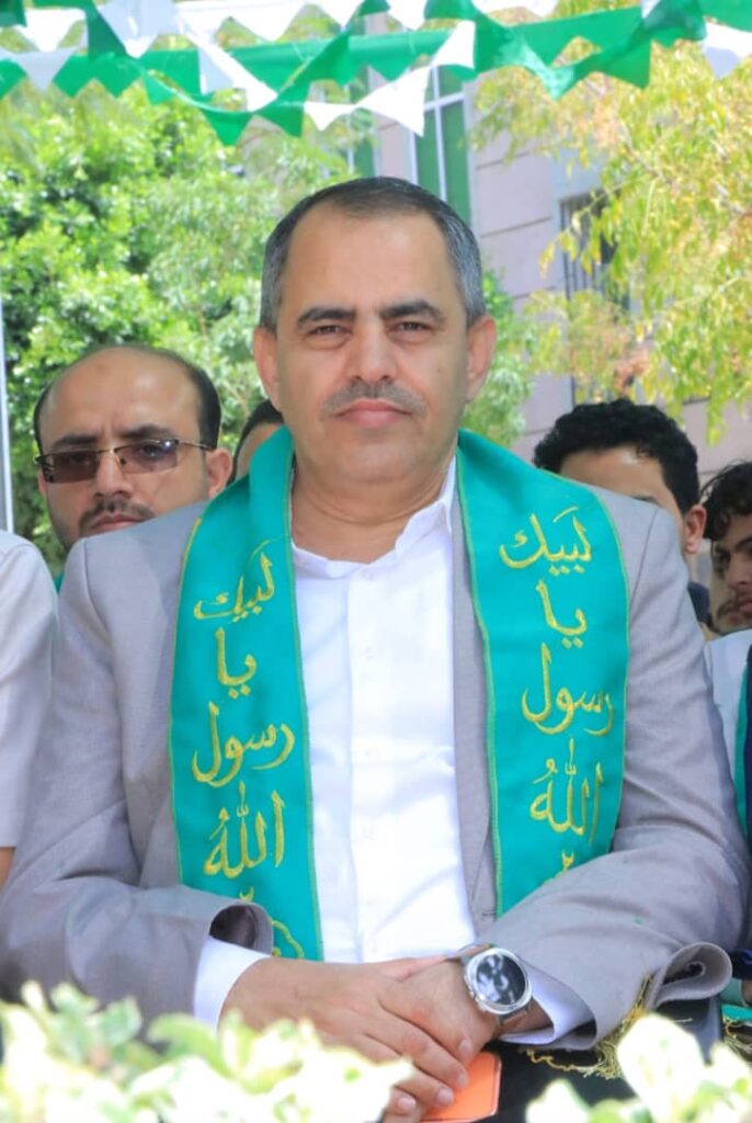Man in a gray suit wearing a green sash with Arabic script at an outdoor ceremony, seated with hands folded.