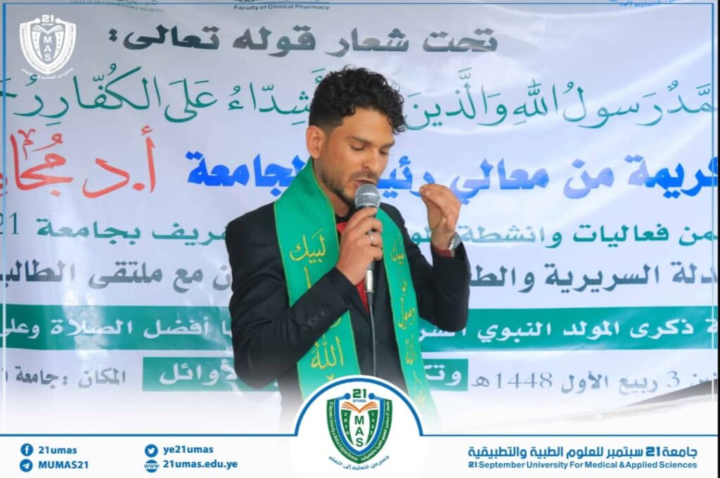 Man in a suit speaks into a microphone at a university event, wearing a green scarf, with a large Arabic banner behind him and a logo at the bottom edge.