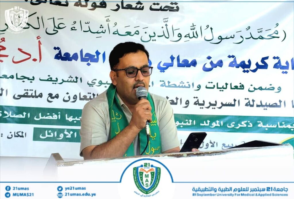 Man with glasses speaks into a microphone at a podium, with a banner containing Arabic text and a university crest visible in the background.