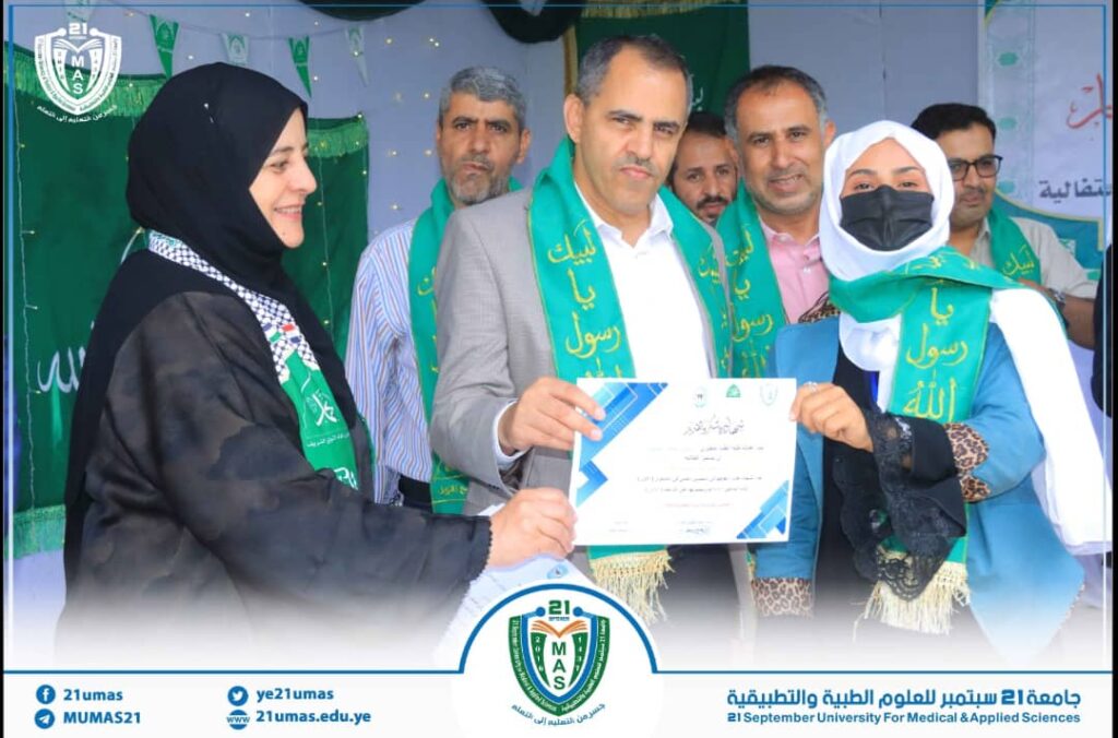 Group of people at a formal ceremony presenting a certificate; two women in hijabs and others wear green sashes with Arabic text; a man in a suit receives the certificate.