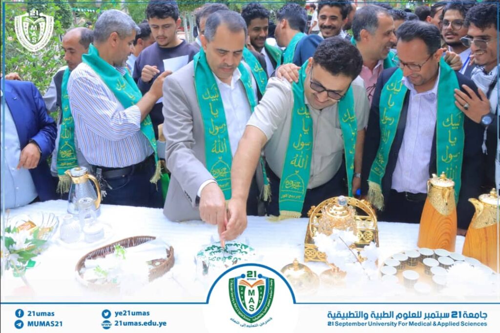Group of men in green sashes cut a cake at a university ceremony, with a blue-and-white banner and the 21 MUMAS logo visible in the foreground.