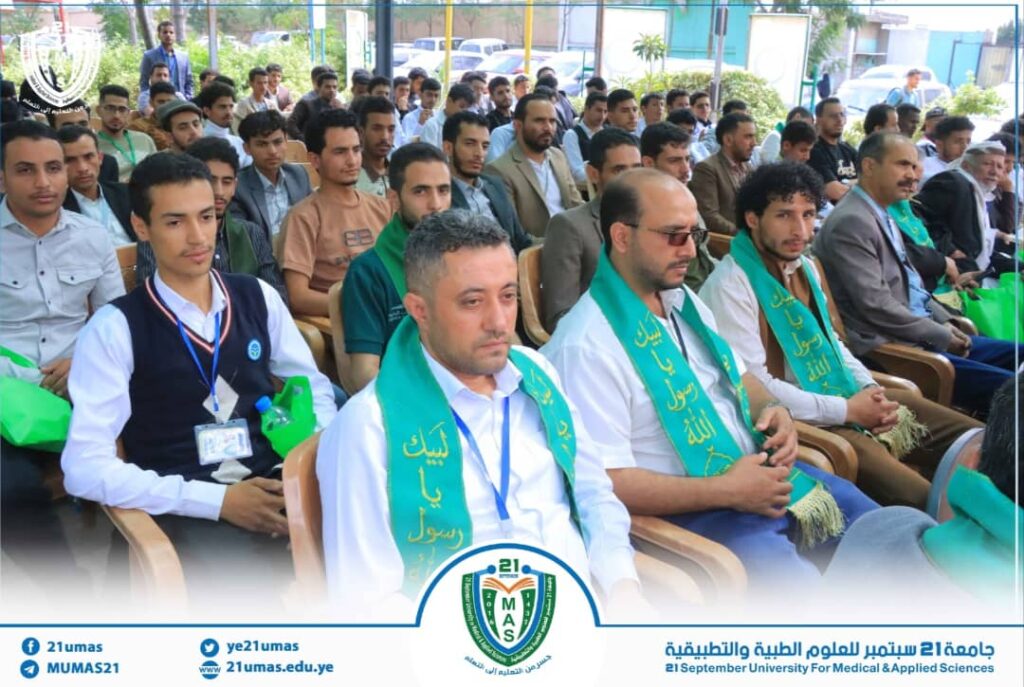Audience of men seated in rows at an outdoor university event, several wearing green sashes across their chests, foreground focused on a few in front row.